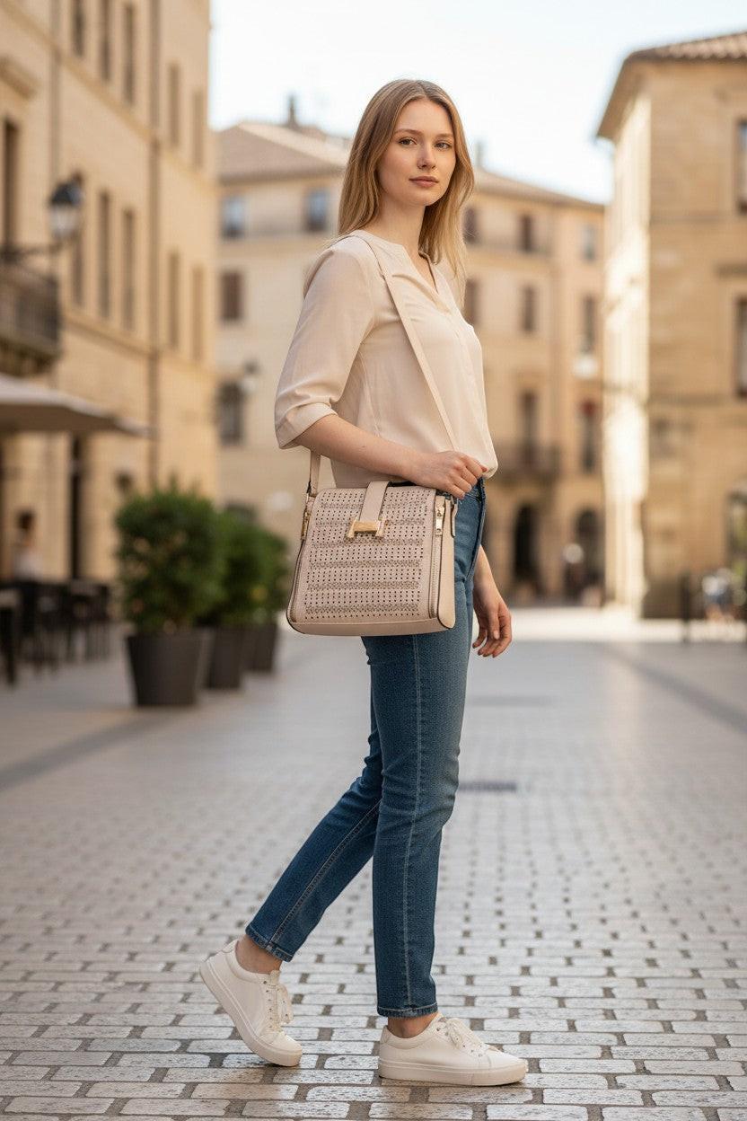 Falio beige faux leather crossbody bag with gold hardware and perforated detail, worn by a woman in jeans and a cream blouse.